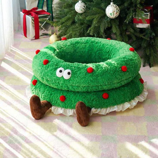 Christmas Wreath Shaped Cat Bed with plush green fabric, red pom-poms, and cartoon eyes, placed under a decorated Christmas tree.
