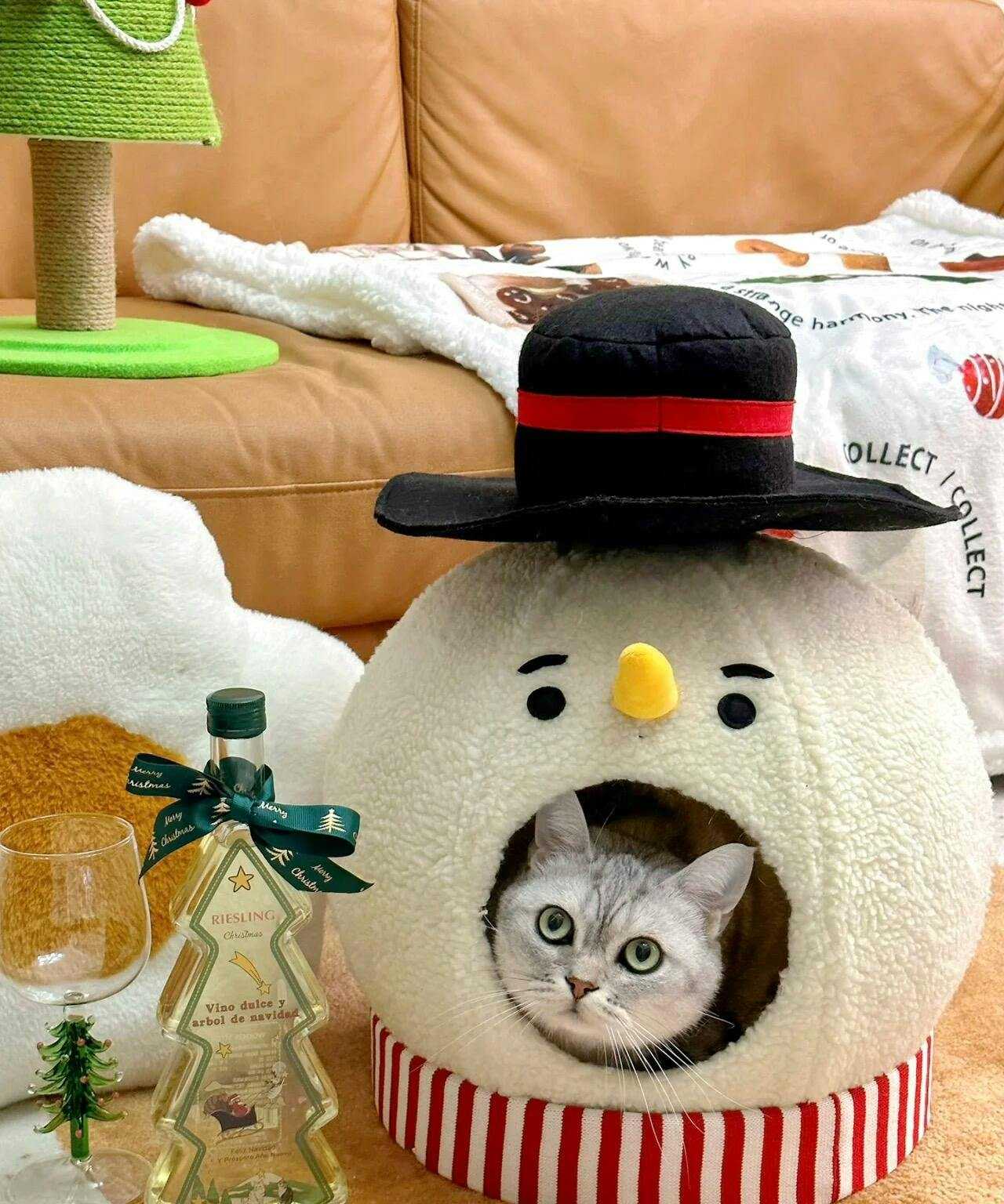 Snowman Cat Covered Bed with cat peeking out, set in cozy living room with wine bottle and festive decor.