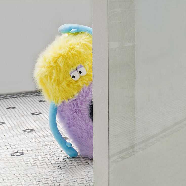 Little Monster Stool Cat Bed peeking from behind a wall, showcasing its quirky monster design with plush texture and blue arms.
