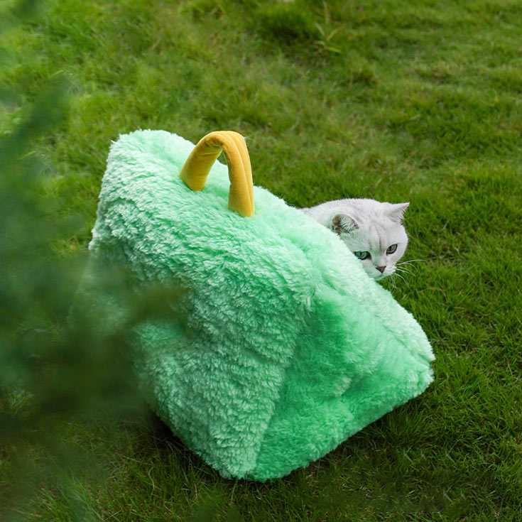 Cat peeking from behind a green Monster Series Cat Bed with yellow handle, showcasing its portable and playful design.