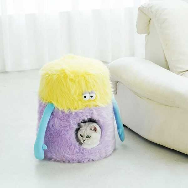 Little Monster Stool Cat Bed in pastel yellow and purple with a cat inside, placed beside a white sofa. Fun cat furniture for cozy lounging.